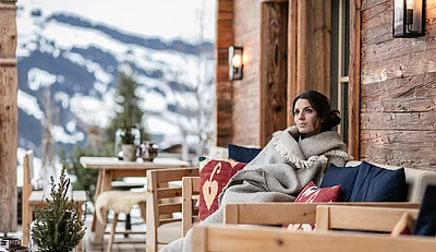 A woman sits snuggled up in a blanket on a cosy bench on the terrace of the luxury mountain chalet Die Schmiedalm