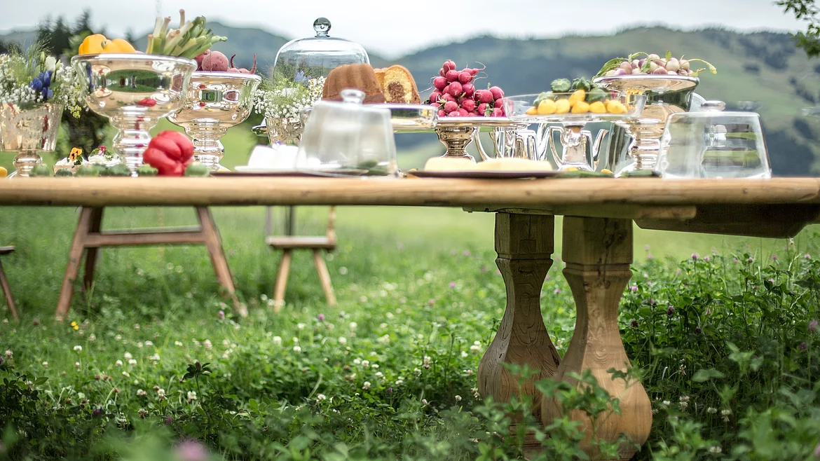 Richly laid wooden table with silver bowls, vegetables and flowers on the meadow in front of the Schmiedalm.