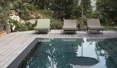 Frau mit Hut lesend auf Sonnenliege am Outdoor-Pool des Chalet Die Schmiedalm in Saalbach Hinterglemm im Sommer mit Chalet und Bergpanorama.