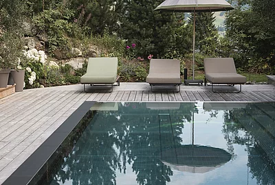 Frau mit Hut lesend auf Sonnenliege am Outdoor-Pool des Chalet Die Schmiedalm in Saalbach Hinterglemm im Sommer mit Chalet und Bergpanorama.